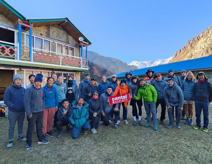 A large group of people in warm outdoor clothing posing together in front of a two-story lodge with wooden balconies in a mountainous region. Snow-capped peaks are visible in the background, and some individuals are holding a red sign that reads 'Cantal Auvergne.' The group appears to be on a trekking or expedition trip